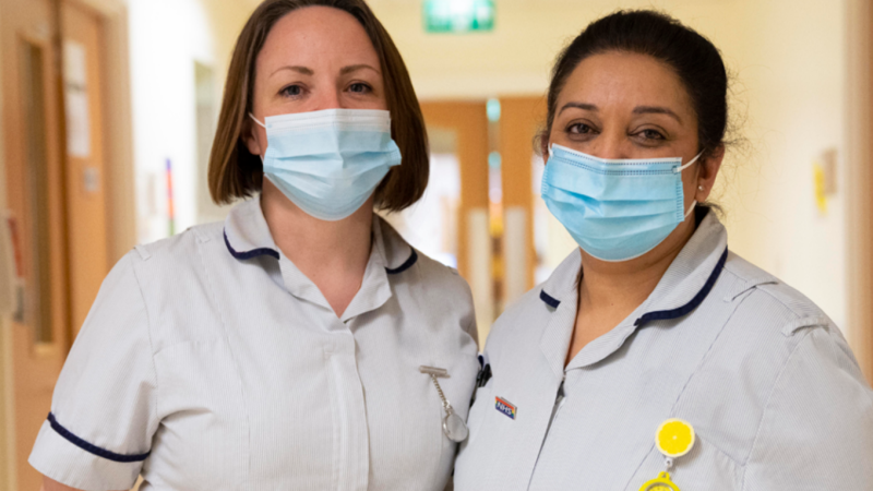 Two nurses wearing face masks standing next to each other in a hospital.