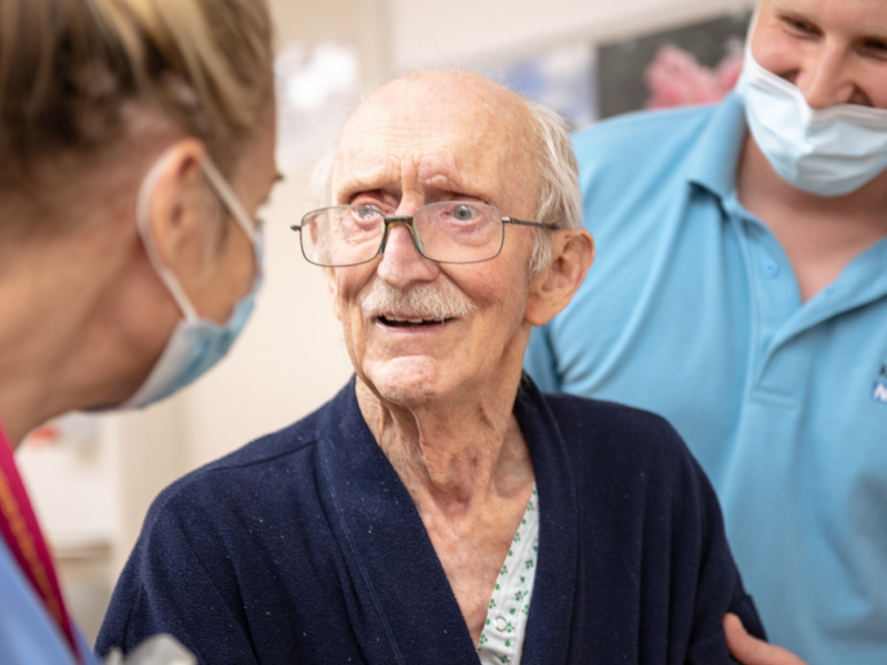 Two female nurses in face masks talking to an older male patient wearing glasses at Kingston Hospital