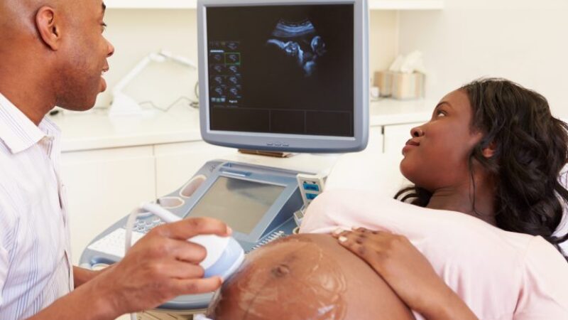 A sonographer performing a scan on a pregnant black woman, who is looking at the scan of her baby on the screen
