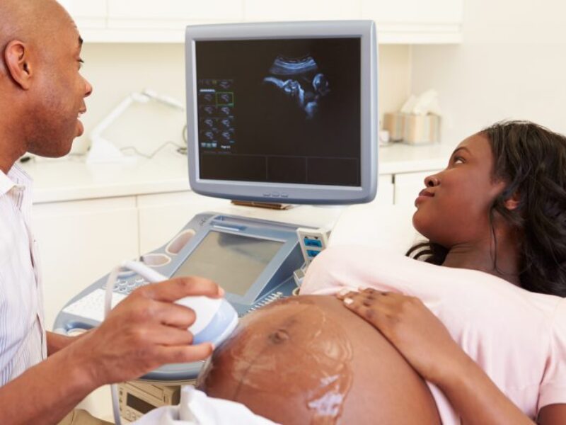 A sonographer performing a scan on a pregnant black woman, who is looking at the scan of her baby on the screen