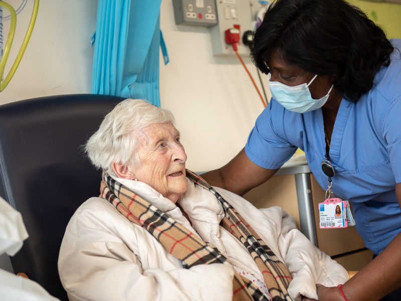 A standing nurse leaning down talking to an older female patient who is sitting in a chair