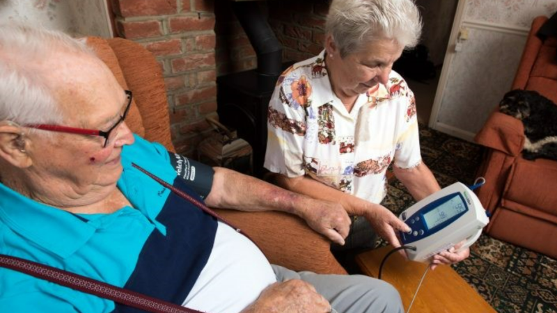 An older woman helps an older man use a remote home blood pressure monitor in their living room.