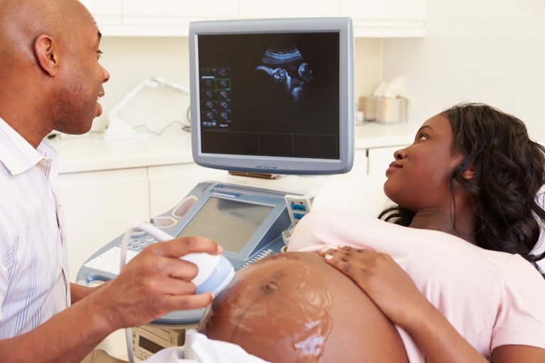 A sonographer performing a scan on a pregnant black woman, who is looking at the scan of her baby on the screen
